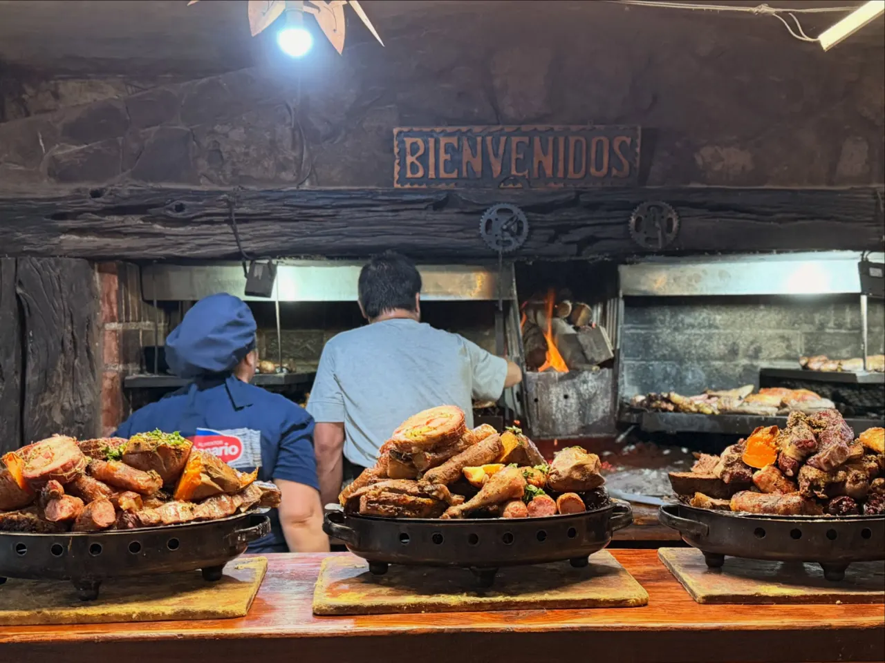 La parrilla de La Bandera con el cartel de madera que dice Bienvenidos, el parrillero atendiendo el fuego y tres braseros cargados de carne y boniato